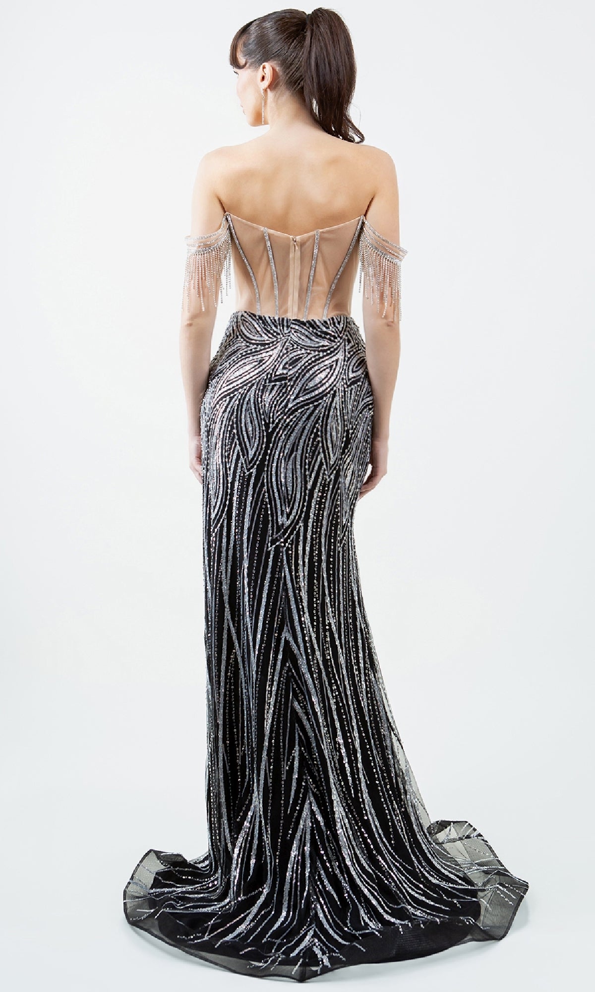 A woman wearing the Cinderella Couture 8147J—a long, off-shoulder black prom dress with silver beaded patterns and a sheer back—stands facing away from the camera.